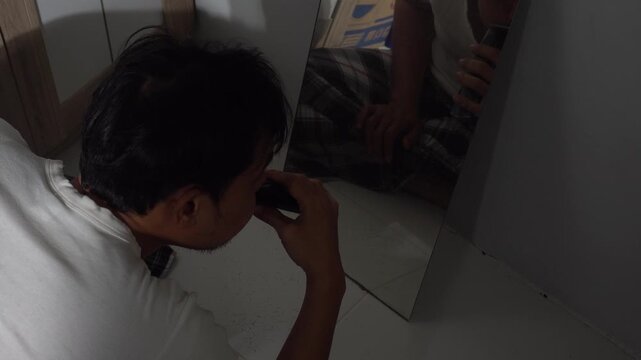 Rear view of an Asian man cutting and grooming his own hair with an electric trimmer clipper in front of a mirror on the floor.