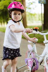 A girl in a helmet with a bicycle walks along a path in a park.