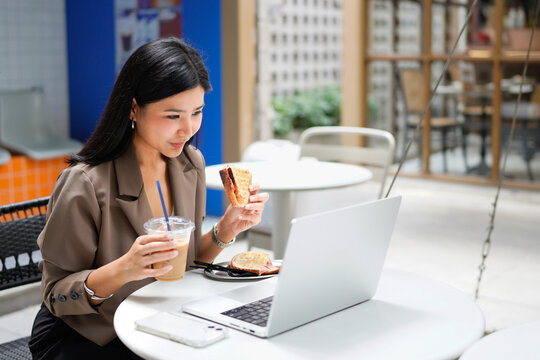 Beautiful Asian businesswoman eating breakfast in a cafe with a sandwich and coffee, and using laptop for work