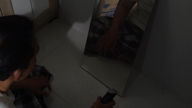 Asian man shaving his beard with an electric trimmer looking at mirror on floor at home. Concept for men grooming routine, hair trimming, personal care, home haircut, daily hygiene, adult male lifesty