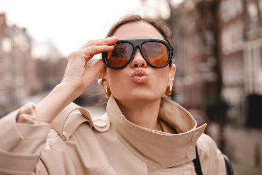 Close-up of a playful young woman in a beige trench coat and large black sunglasses posing with a kissy face on a blurred city background with historic architecture and cozy autumn street light