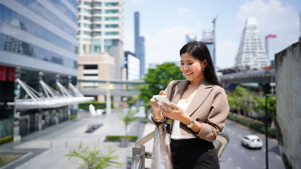 Fototapeta premium Successful Asian businesswoman smiling while using smartphone standing on balcony surrounded by modern skyscrapers in the urban financial district