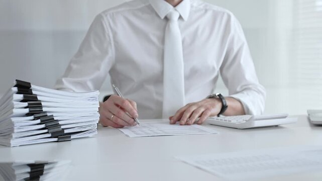 Businessman working at a desk, reviewing official documents, writing with a pen, and using a calculator, managing a stack of organized paperwork in a professional office environment. Audit and taxes