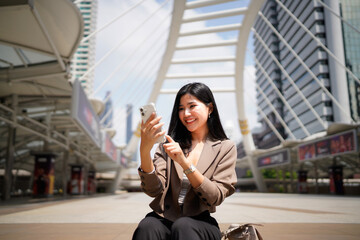 Obraz premium Successful Asian businesswoman smiling while using smartphone sitting on stair surrounded by modern skyscrapers in the urban financial district
