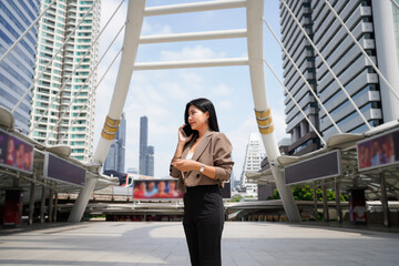 Fototapeta premium Asian businesswoman talking on mobile phone walking in city street to office, skyscrapers cityscape background. Young woman on smartphone happy, woman making business call on cell