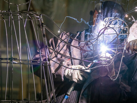 Close-up of a professional metalworker in a protective welding mask and heavy-duty gloves using a welding torch on a complex steel wire structure, with bright electric arcs