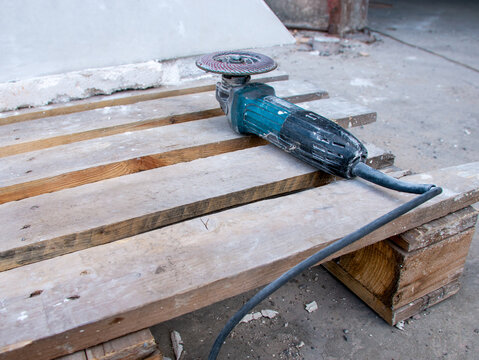 Professional blue handheld electric angle grinder with abrasive flap disc resting on rough wooden pallet inside dusty industrial workshop construction environment building equipment.