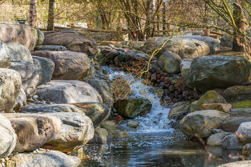 Rustic stream cascades over mossy stones. Bark and branches frame the natural flow. Sunlight glints on moving water and wet rock. Peaceful, quiet atmosphere invites calm reflection © Jakob