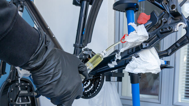Hands in gloves are performing bicycle hydraulic disc brake bleeding. A syringe with yellow fluid connects to the caliper, flushing the system
