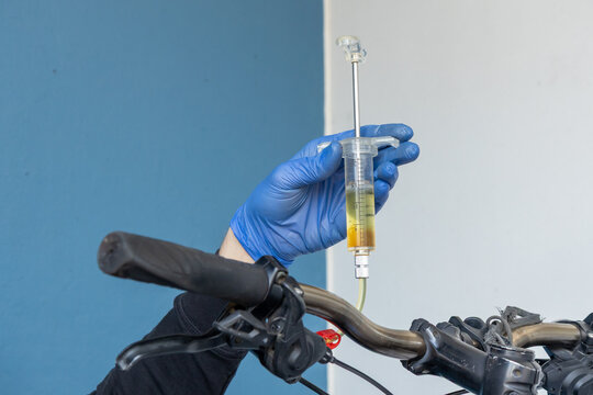 A professional bicycle mechanic wearing blue protective nitrile gloves uses a specialized syringe kit to perform a hydraulic disc brake bleed on a mountain bike handlebar in a repair workshop