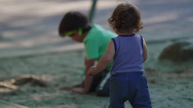 Toddler rising from sand with determined posture and walking forward in sandbox holding small bucket capturing persistence balance and confident outdoor childhood exploration