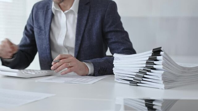 Businessman sorting through stacks of documents on a white desk beside a tall pile of bound papers, symbolizing meticulous financial record keeping and bureaucracy. Audit and taxes