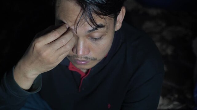 Top view of a stressed Asian man looking down with a sad expression, touching his forehead and hair in distress. Concept of depression, anxiety, headache, or mental exhaustion. Moody lighting emphasiz
