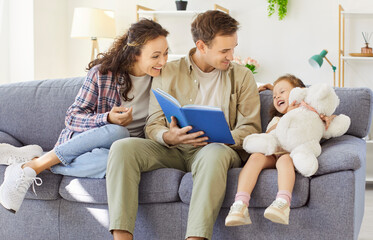 Happy parents and little child daughter reading book together. On a cozy sofa at home, they smile,...