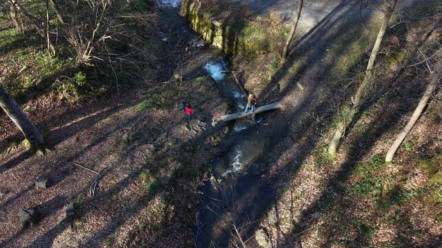 Man standing on a wooden plank over a stream throwing a stone into the water while children watch nearby. Natural outdoor family activity, exploration and connection with nature.