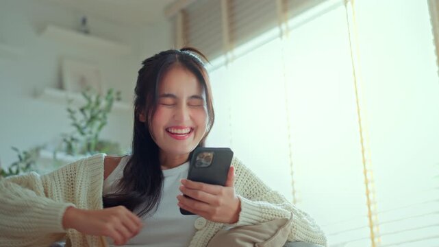 Excited young Asian woman celebrating success while looking at her smartphone at home, raising her fist in a winning gesture, depicting digital achievement, good news, and happy emotions in a bright