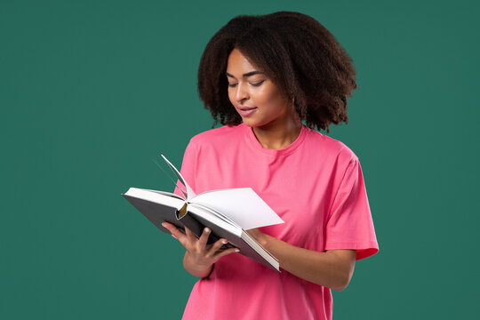 Pretty reader woman stands against green backdrop, holding and reading book with curious, engaged expression. Smart girl. Discovery, learning, intellectual curiosity, spending quality time