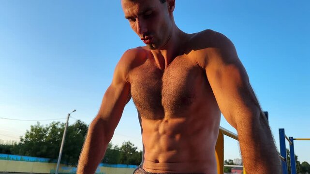 Group of young shirtless sportsman trains abdominal muscles raising legs on parallel bars at sports ground. Strong man exercising at nature. Sporty athlete working out at nature. Sportive lifestyle