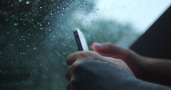 Tattooed Hand Using or Holding Smartphone By Rain-Covered Car Window, Soft Focus Wet Glass, Moody Travel Atmosphere, Mobile Use During Stormy Weather