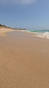 A low-angle vertical view of a pristine coastline with turquoise waves crashing on golden sand, dunes, and blue sky. The wild and famous beach is called Bolonia, in Cadiz, Andalusia, Spain