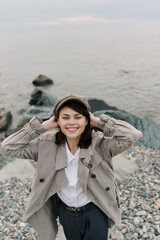 Smiling young woman in a hat and patterned coat enjoys a pebble beach with calm water in the...