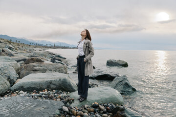 Young woman stands on rocky shore near calm sea under cloudy sky, wearing casual clothes. Concept...
