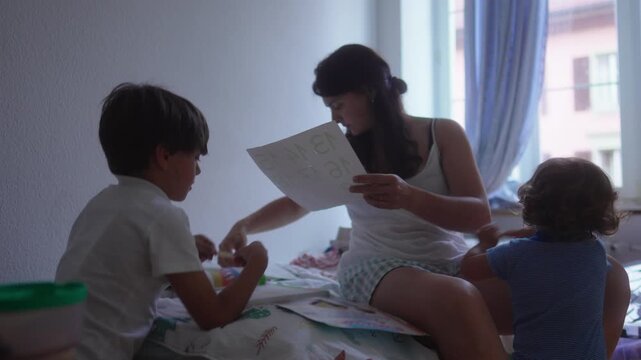 Mother sitting on bed holding educational number sheets while children gather around during home learning activity, family bonding, curiosity, early education and playful discovery
