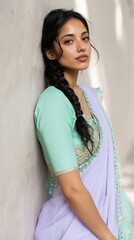 Fototapeta premium Adult asian female with long braided hair wearing mint green blouse and lavender sari leaning against wall soft natural light confident look
