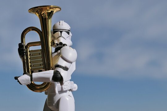 Action figure of Star Wars clone trooper in white helmet and armor holding model of some kind of euphonium brass instrument, blue spring skies with overcast clouds in background. 