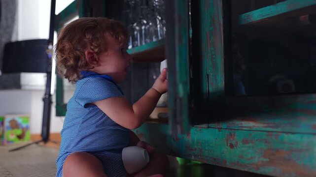 Toddler sitting beside wooden cabinet taking small cups from shelf while exploring household objects during playful moment of curiosity discovery and toddler mischief at home