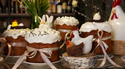 Easter cakes decorated with white glaze and satin ribbons near vintage figures of a hen and a cockerel close-up against the background of shelves as a décor for a spring holiday © Irina