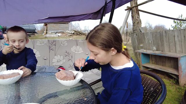 Two children enjoy ice cream while sitting at a glass table in the backyard. They are relaxed, focused on their treat, with a few distant objects visible in the background.