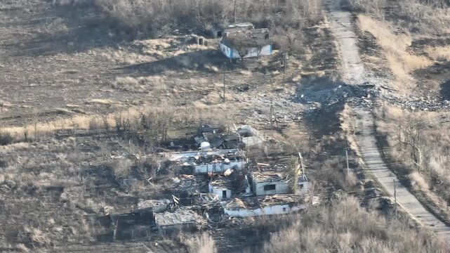 Aerial drone footage captures a heavily damaged building with a collapsed roof and debris, surrounded by craters and destroyed structures in a conflict zone.