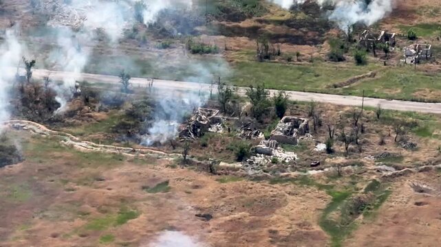 Aerial drone footage captures the aftermath of a military strike on a rural settlement. Thick white smoke rises from multiple impact points near a paved road and destroyed buildings. The landscape is 