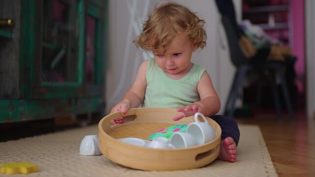 Toddler sitting on floor sorting small cups and plates from wooden tray focused childhood moment of exploration organization and tactile play with household objects at home