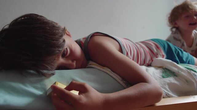 Young boy lying on bed holding small object with distant gaze expressing boredom and disengagement while sibling plays in background during quiet childhood moment at home