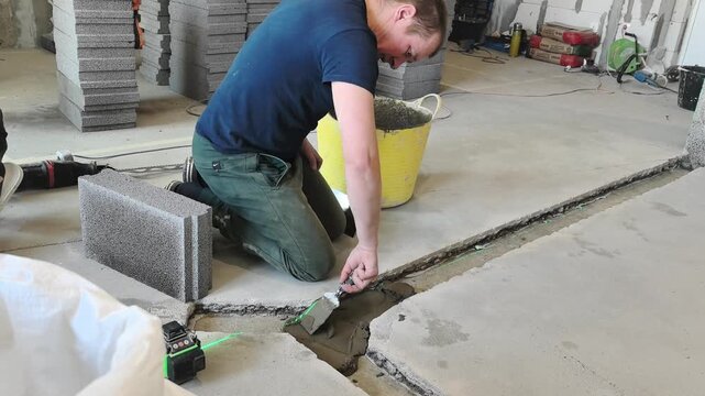 A man works on fixing a concrete floor in a construction area. He uses a trowel to fill cracks and ensures proper application of materials. The space is busy with ongoing tasks.