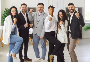 Group of young multiethnic teammates stands proudly in office, looking at camera and raised fists...