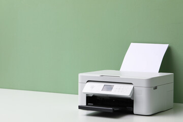Fototapeta premium One printer with sheet of paper on white table near green wall, closeup. Space for text