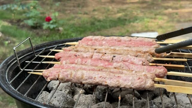 Skewers of minced meat cook on a charcoal grill outdoors, smoke and heat suggest a casual backyard barbecue, warm appetizing mood for summer gatherings