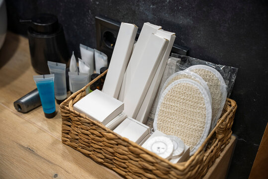 Luxury hotel bathroom amenities set in a woven basket on a wooden countertop. Includes white branded boxes, loofah sponges, shower gel, and toiletries against a dark slate wall. Minimalist spa.