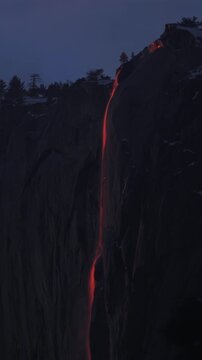 Vertical Screen: Yosemite National Park Firefall phenomenon creates stunning orange glow cascading down steep granite rock face at twilight with silhouetted pine trees above