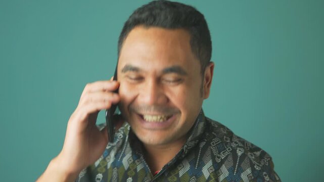Cheerful Asian man in a batik shirt talking on a mobile phone, laughing heartily and looking away. Concept of good news and happiness.
