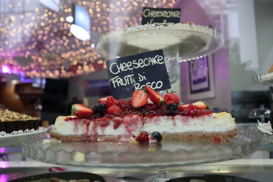 Cheesecake ai frutti di bosco esposta su alzata in vetro, con strato cremoso e topping di fragole, mirtilli e lamponi, fotografata in un ambiente di caffetteria per esaltare colori, texture e presenta