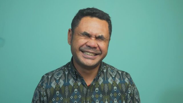 A friendly Asian man in a traditional batik shirt smiling broadly and genuinely at the camera against a teal wall. Concept of happiness and success.
