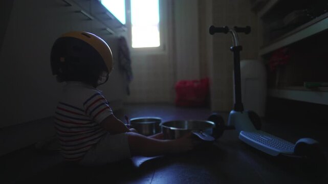 Toddler sitting on kitchen floor with helmet tapping metal pots beside scooter showing curiosity exploration and playful interaction with everyday household objects at home