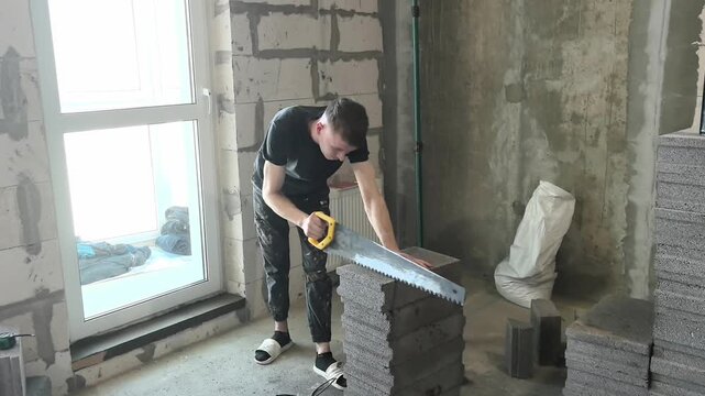 A person cuts building blocks in an empty room. They stand near a large window and work on the construction. Tools and materials are around them, showing the progress of the work.