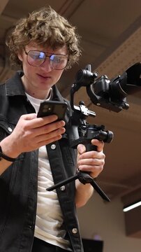 Vertical screen Videographer studies script on smartphone holding camera on gimbal looking confused struggling to understand how to execute idea planning scene in studio