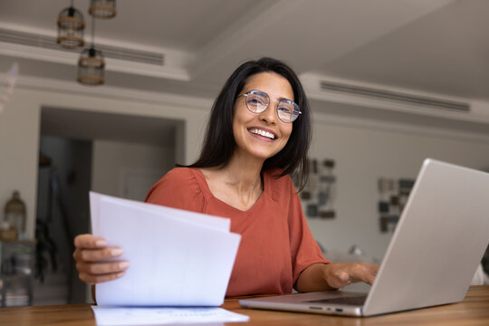 Happy woman in glasses working on laptop, holding documents, busy in financial planning, reviewing family incomes and expenses, preparing tax reports, sitting at table in cozy home interior, portrait