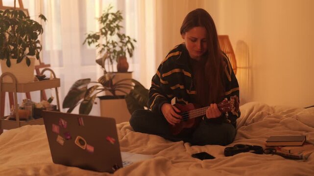 Full view of a young woman sitting cross-legged on a bed, interacting with a laptop before starting to play a ukulele. Warm sunlight creates a cozy, focused, and creative home scene.